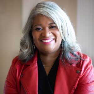 Dark skinned woman with long grey hair smiling and wearing a red blazer and black shirt.