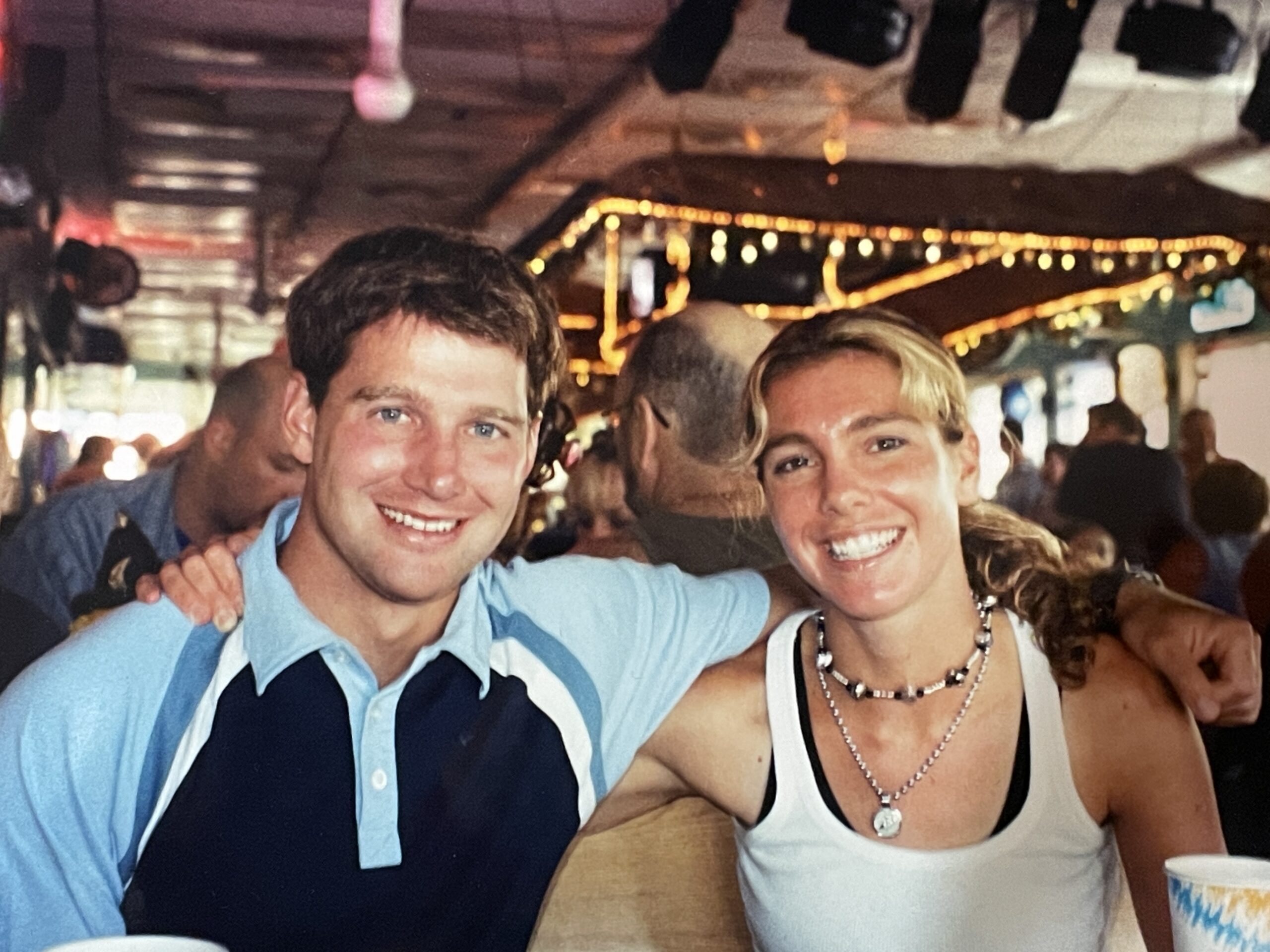 A man wearing a light blue and black shirt, with short dark hair, smiling and a woman wearing a white tank top, with blond hair, smiling. Both are close together hugging and facing the camera.