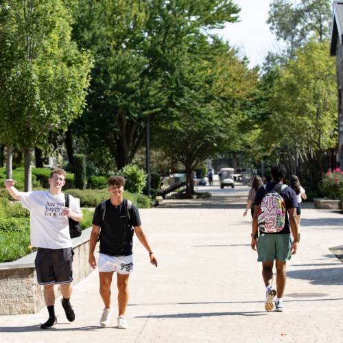 Two men walking on campus. One with a white t-shirt and dark shorts and one with a dark t-shirt and light shorts. Another person walking with a colorful backpack on.